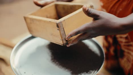 african lady use handmade sifter to sieve dark brown roasted ugandan coffee