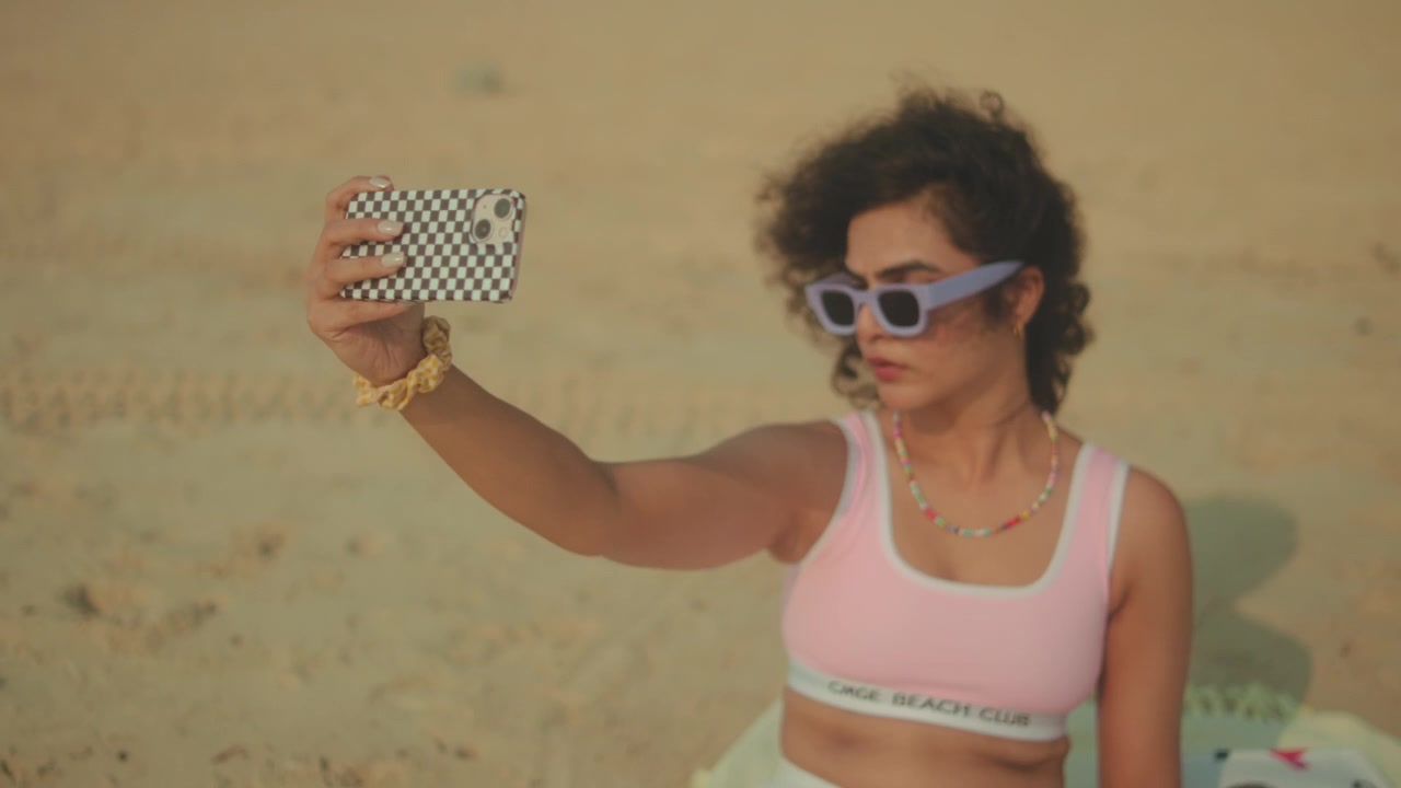 Donna che si fa un selfie con lo smartphone su una spiaggia sabbiosa mentre si gode una giornata di sole all'aperto. Concetto di stile di vita moderno, tra viaggi e social media, che include svago, comunicazione e ricordi delle vacanze.