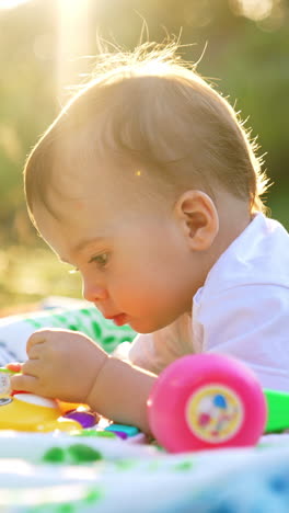 nice little caucasian toddler lies on his belly on the plaid in the garden. lovely child looking at toys in the rays of bright sun. vertical video