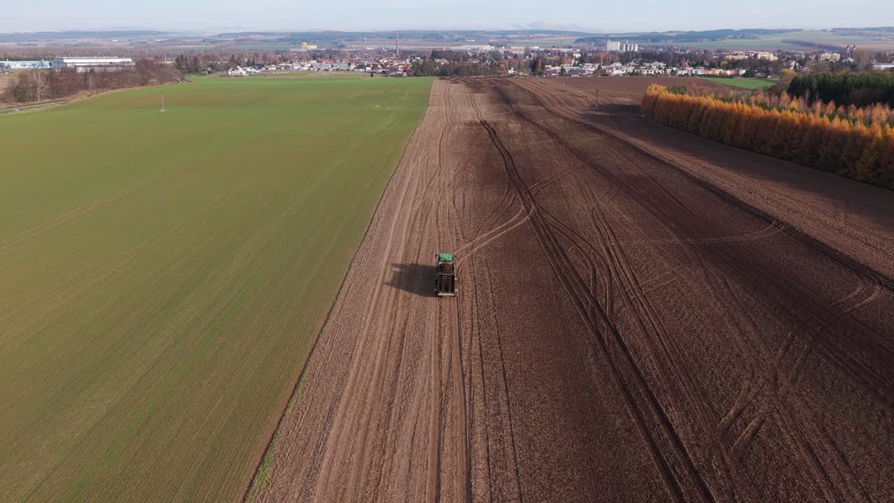 Traktor pflügt und düngt Herbstfelder, aufgenommen von einer Drohne mit einer fernen Stadt am Horizont