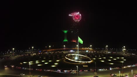 Antena-Circulando-La-Bandera-De-Arabia-Saudita-Ondeando,-Noche-De-Fuegos-Artificiales,-Torre-Khobar-En-El-Fondo