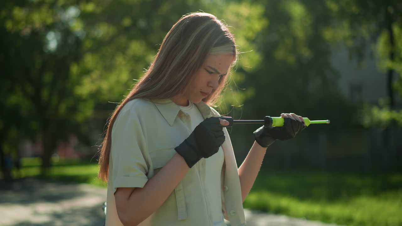Blonde-haired girl standing outdoors with a thoughtful expression, carefully inspecting green air pump handle, softly blurred background of trees and a building