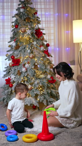 cute baby boy and his mom sit on the floor talking. child and his mother play with a pyramid near the christmas tree. vertical video.