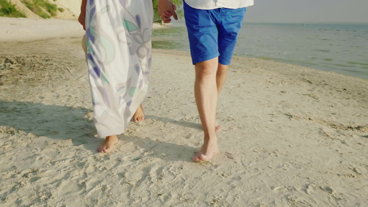 한 젊은 부부가 다리만 보이는 프레임의 해변 모래 위를 맨발로 걷고 있다 v