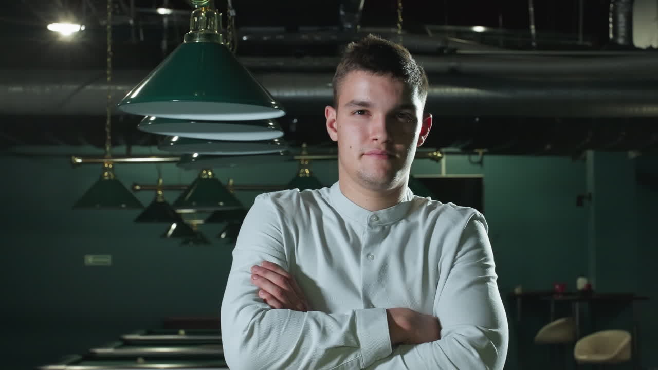 백색 셔츠를 입고 팔을 접고 은 미소를 짓는 신사의 클로즈업. 여러 개의 초록색 술집 테이블, 오버헤드 램프, 현대적인 인테리어는 전문적이지만 편안한 분위기를 만니다.