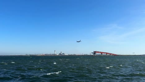 Amplia-Vista-De-Un-Avión-Volando-Sobre-Una-Gran-Masa-De-Agua-Cerca-De-Un-Puente-Rojo-Y-Un-Paisaje-Urbano-Industrial