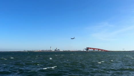 wide view of an airplane flying over a large body of water near a red bridge and industrial cityscape