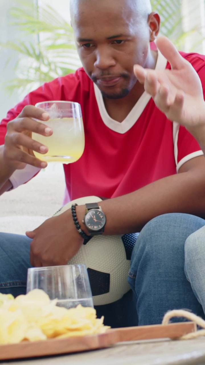 Vertical video: Pan showing man sipping cup in red shirt at home with soccer ball watching match