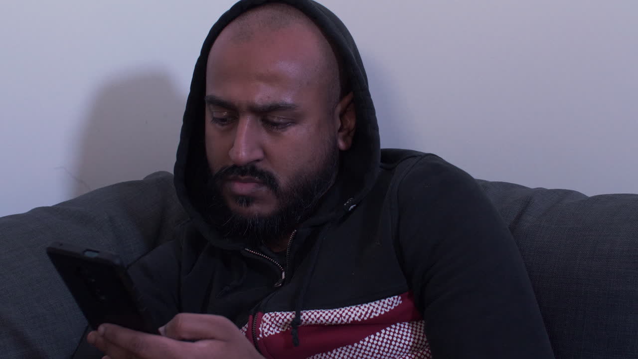 hombre encapuchado en el sofá desplazándose en el teléfono móvil por la noche