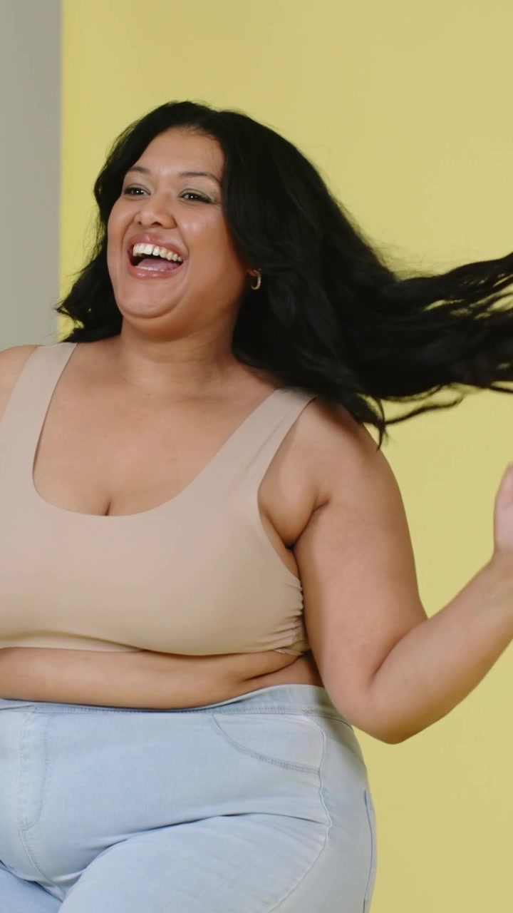 Video verticale: Donna sorridente che si sistema i capelli su uno sgabello per un servizio fotografico, con sfondo giallo e top beige.