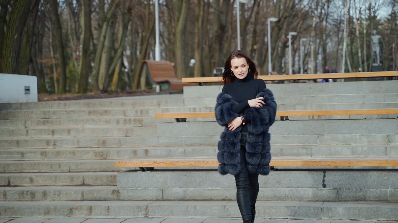 Beautiful model posing on camera in autumn park. Young woman in black fur coat walking and looking at camera. Slow motion.