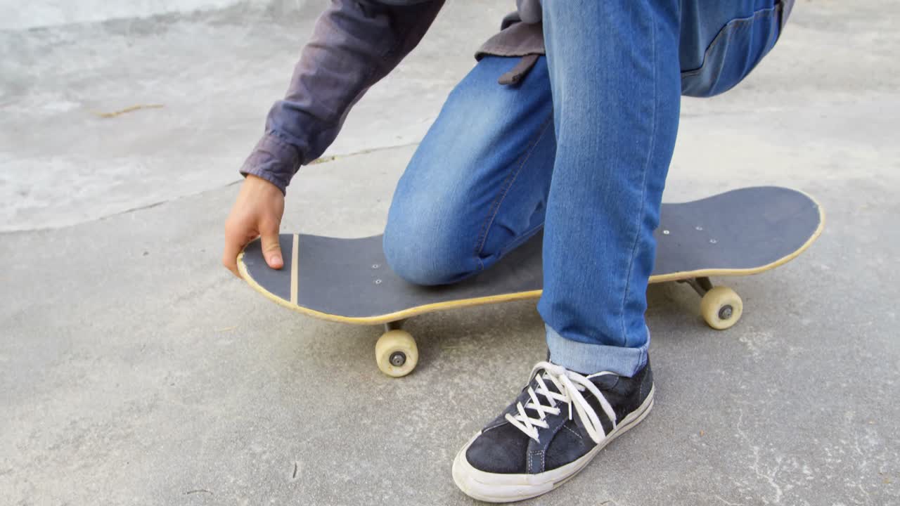Bagian bawah tubuh pria muda mengikat tali sepatunya sambil berjongkok di atas skateboard di taman skateboard 4k.