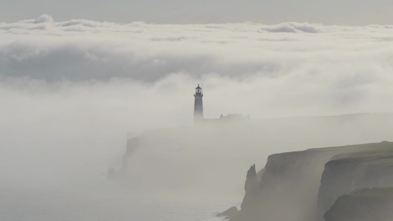 Nebbia bassa che si ritira, rivelando la torre di un faro e scogliere rocciose su un promontorio costiero, con un banco di nuvole.