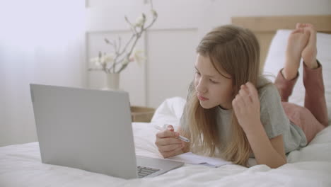 Una-Alegre-Alumna-Está-Respondiendo-Durante-Una-Lección-En-Línea-Con-Una-Computadora-Portátil-Acostada-En-La-Cama-Y-Hablando-De-Aprendizaje-Electrónico