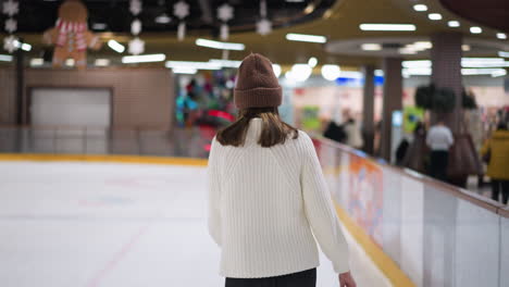 a close view of a lady skating slowly on an ice rink, wearing a white sweater and brown beanie, with people standing by in the background