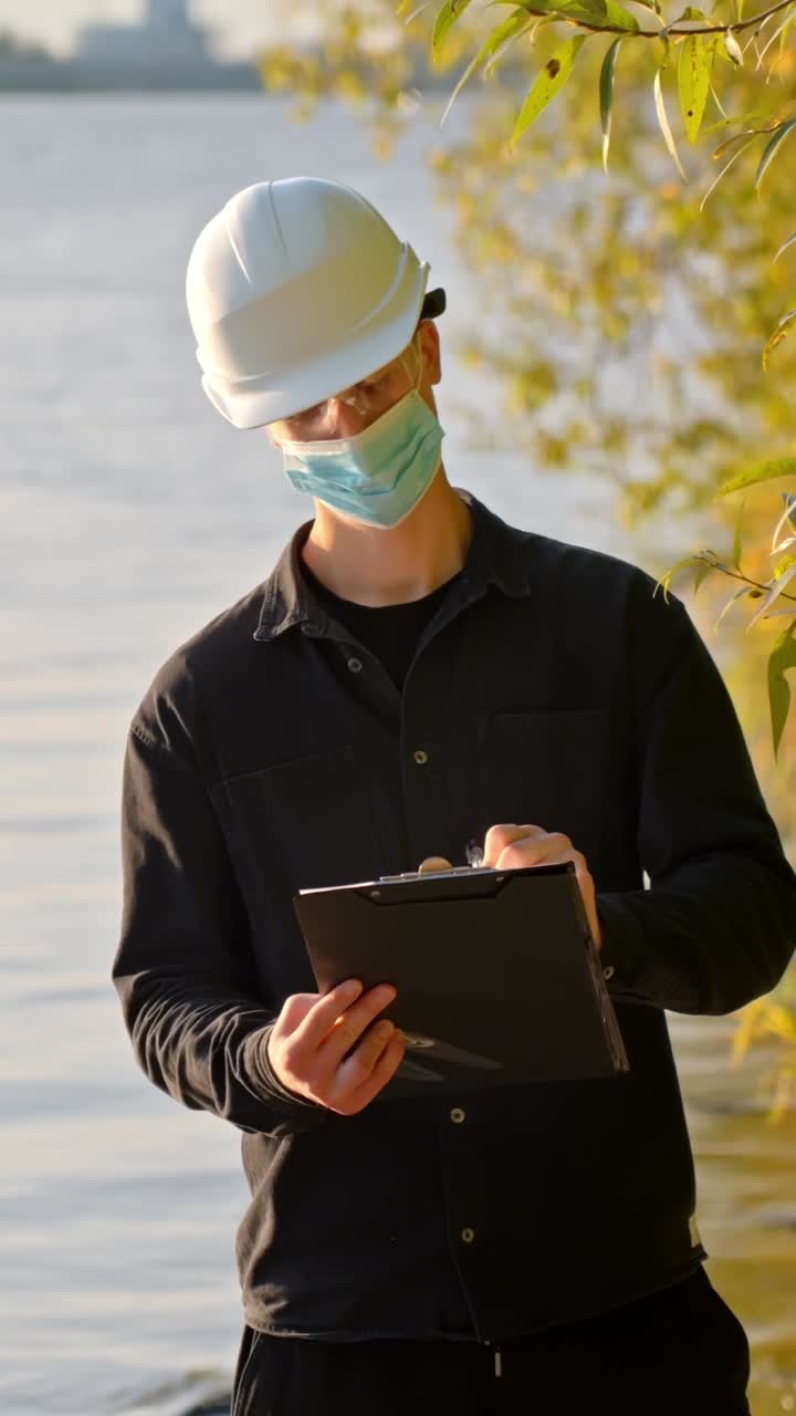 Masked person stands near stream looking up at infected plant taking notes on clipboard, vertical