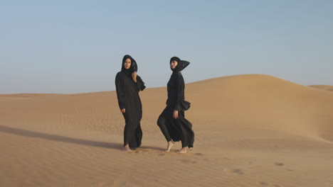 Zoom-In-Aufnahme-Von-Zwei-Schönen-Muslimischen-Frauen-Im-Hijab,-Die-In-Einer-Windigen-Wüste-Posieren-Und-In-Die-Kamera-Schauen