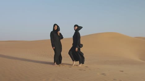 zoom in shot of two beautiful muslim women in hijab posing in a windy desert and looking at camera