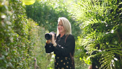 blonde photographer with dslr camera posing subjects for outdoor photo shoot in green garden, 4k resolution slow motion