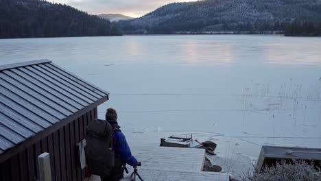 Backpacker-Man-Accompanied-By-His-Alaskan-Malamute-With-Vast-Frozen-Lake-In-Coniferous-Woodland-Hills-Near-Trondheim,-Norway