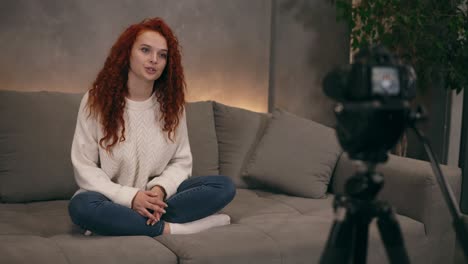 Curly-redhead-young-girl-vlogger-is-talking-in-front-of-camera-recording-video-for-online-blog-in-internet-speaking,-smiling