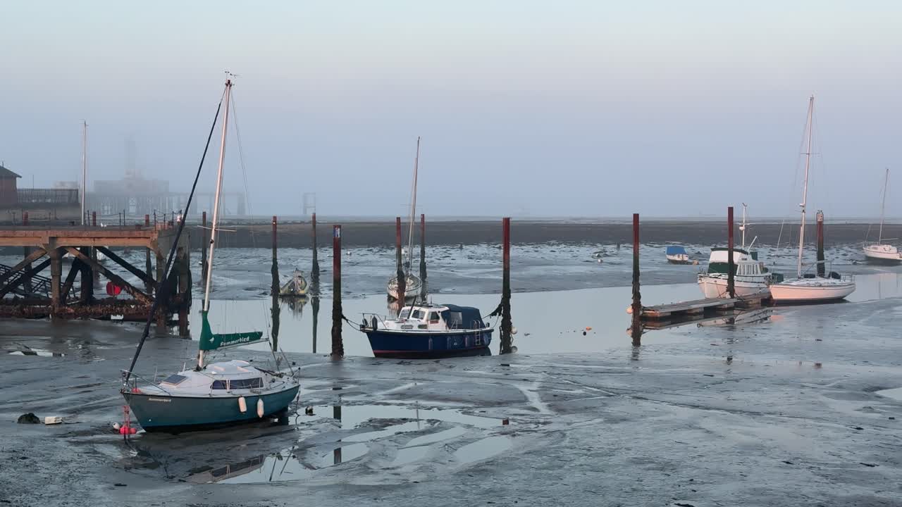 En panorering afslører både og sejlbåde fortøjet i mudderflader ved lavvande en tåget morgen i Gosport, England, med en mole og bygninger i baggrunden.