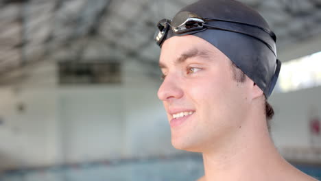 smiling male swimmer wearing swim cap and goggles, standing by indoor pool, copy space