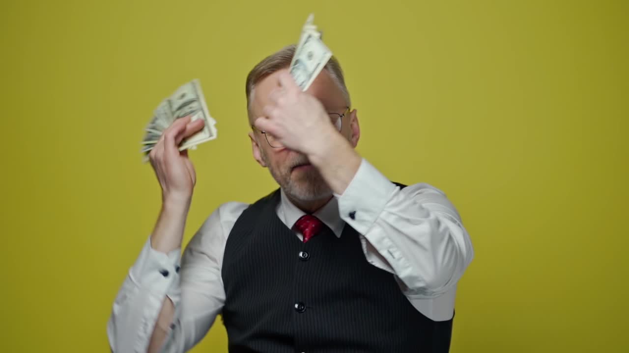 Excited mature man with dollars in hands. Happy elderly man is waving many dollar bills isolated on yellow background. Slow motion. Positive emotions.