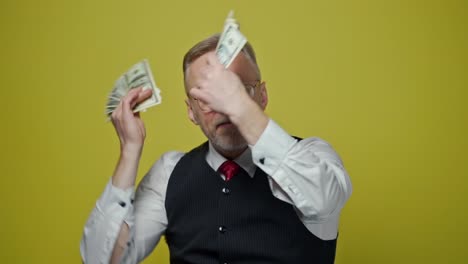 excited mature man with dollars in hands. happy elderly man is waving many dollar bills isolated on yellow background. slow motion. positive emotions.