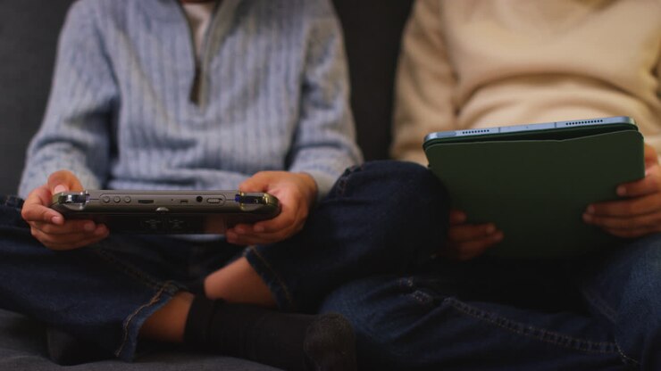 Nahaufnahme von zwei jungen Jungen, die zu Hause auf dem Sofa sitzen und Spiele auf einem digitalen Tablet und einem Handheld-Gaming-Gerät spielen oder streamen