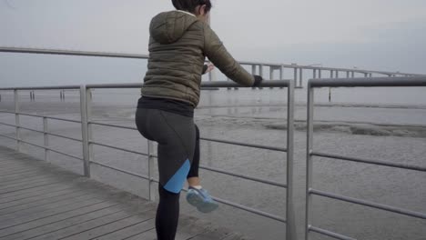 Slow-motion-shot-of-concentrated-sporty-young-woman-stretching-legs-on-wooden-jetty-with-metal-fence