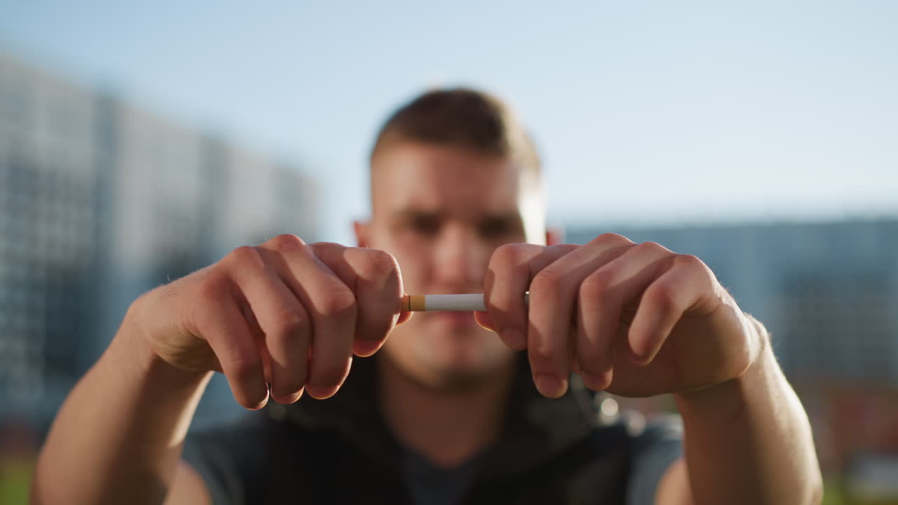 boy extends hands forward gripping cigarette and snaps it in half with determined expression, skin highlighted by sunlight, urban brick building and blurred buildings under bright daylight