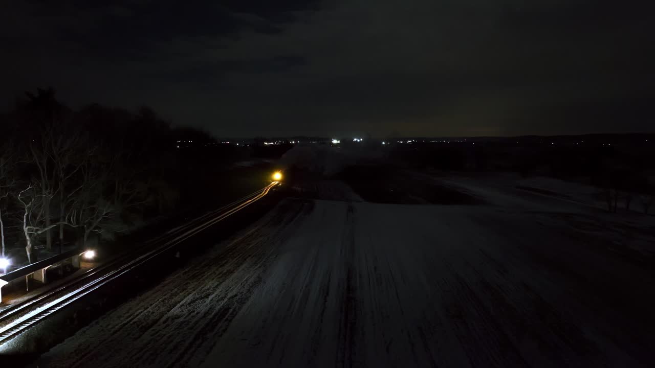 A train moves through a snow-covered rural area under a dark sky. Bright lights from the surrounding landscape and city create a striking contrast against the night backdrop.
