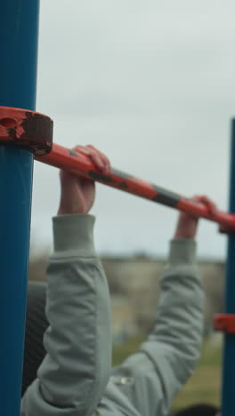 Two boys are working out on pull-up bars in an outdoor gym area, each gripping the bars but appearing tired and struggling to continue their exercises
