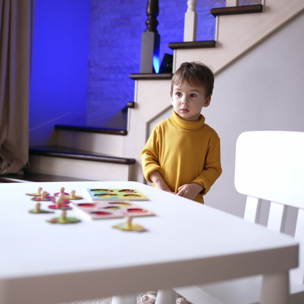 Bambino con un maglione giallo sta in piedi vicino alla scrivania. Il bimbo volge la testa verso la finestra, guardando fuori.