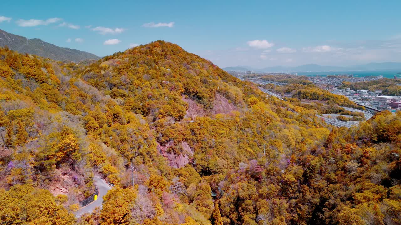 A slow aerial pull-out shot reveals a vast Japanese mountain landscape covered in vibrant, golden autumn foliage, with a winding road and distant coastal city.