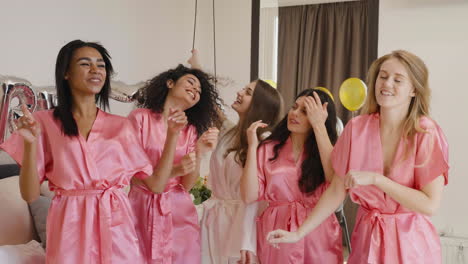 grupo de amigas multiétnicas y novias con camisones de seda rosa y blanca mientras bailan en una habitación en una reunión nupcial 1