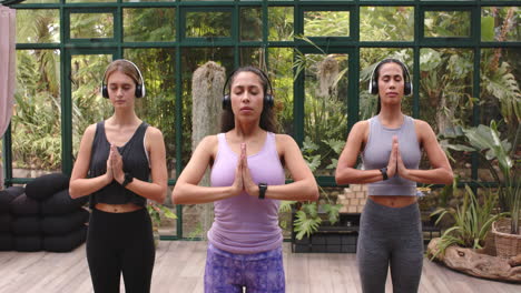 practicing yoga, three women in activewear meditating with headphones in glass house