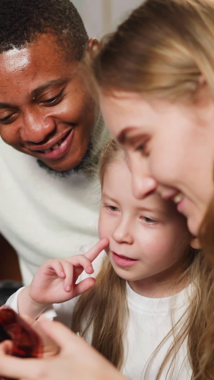 Happy African-American man with daughter and white wife looks at processed coloured film sitting on sofa at home close view slow motion
