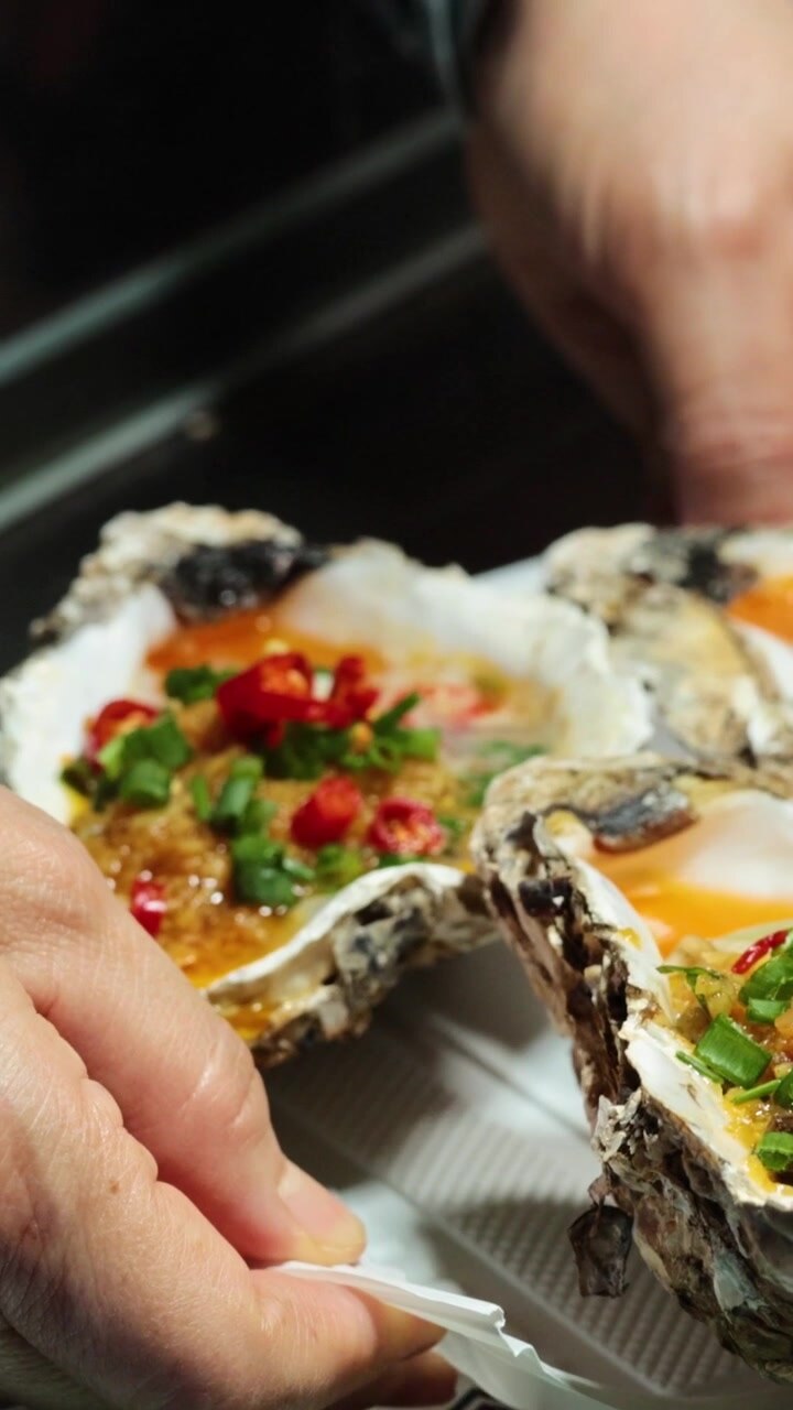 A close-up vertical shot of hands holding a tray of oysters topped with chili.