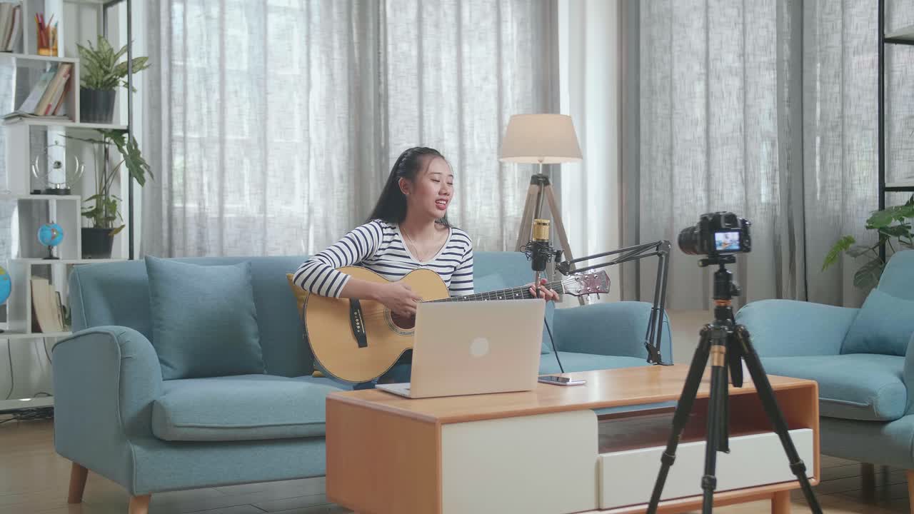 mujer asiática con una computadora portátil cantando y tocando una guitarra mientras transmite en vivo en casa