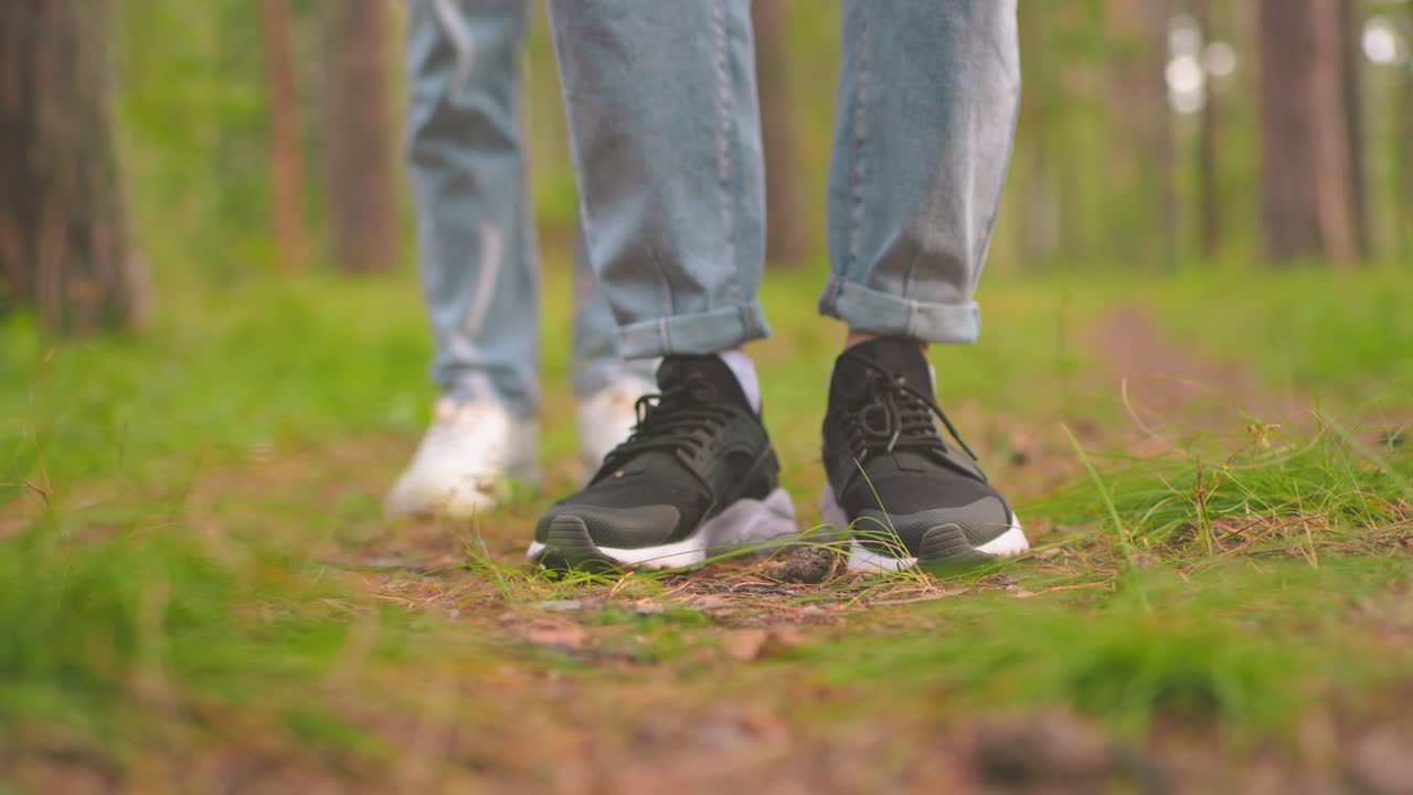 Close-up of two individuals standing on forest trail in blue jeans, one in black sneakers and other in white sneakers, person in black sneakers taps leg on the ground