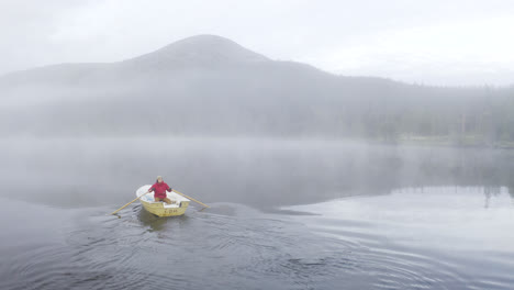 hombre en una chaqueta roja remando un bote pequeño en un lago lleno de niebla