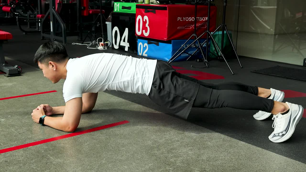 Athletic man holds forearm plank in modern gym, side view, surrounded by colorful equipment