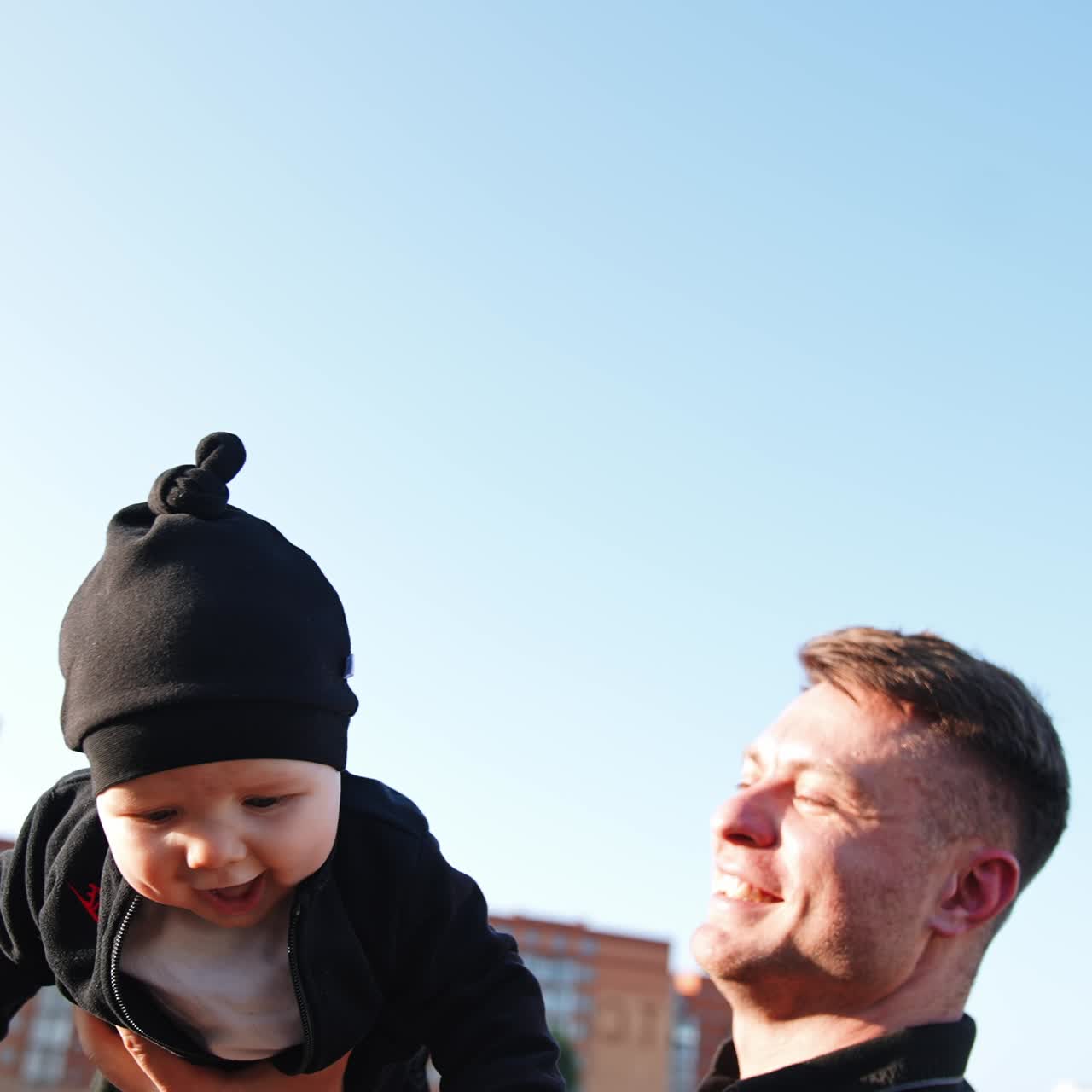 Uomo sorridente solleva il suo bambino sopra la testa con una mano. Simpatico bambino sorride in modo adorabile. Cielo azzurro sullo sfondo. Angolazione dal basso