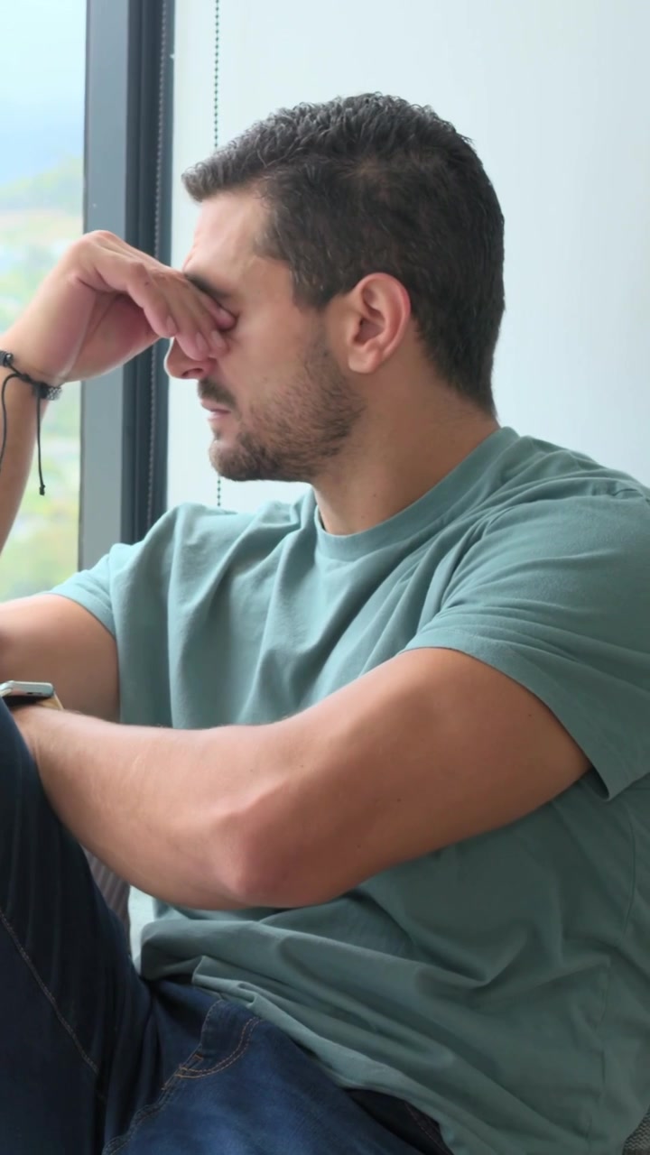 Video verticale: Uomo con maglietta verde acqua che reagisce alla vista esterna, strofinandosi il naso e scrutando la finestra a casa.
