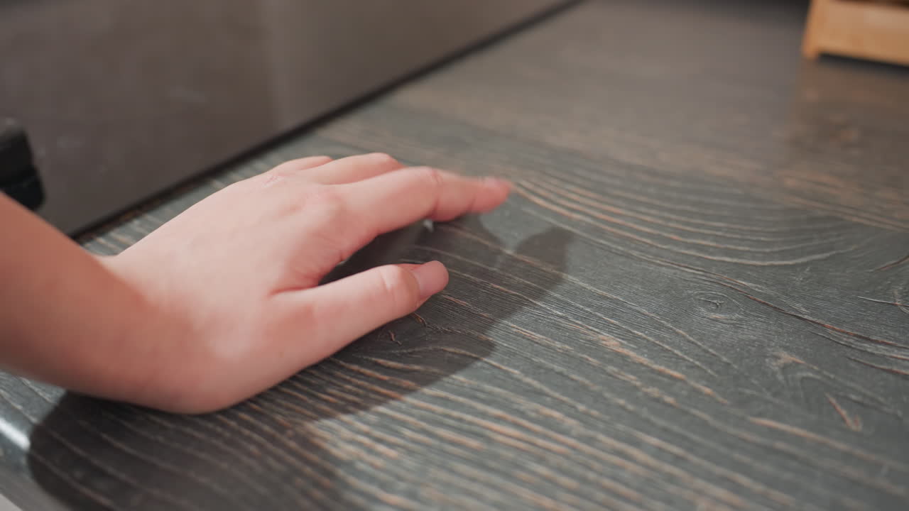 Primo piano della mano di una donna con unghia curata che tamburella delicatamente le dita sul piano di lavoro della cucina in legno scuro, accanto a un fornello nero, in un interno domestico moderno, esprimendo attesa.