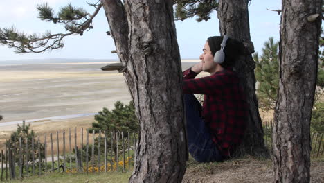 shot of an authentic hipster man listening to music with headphones whilst sitting in the sun against a tree trunk