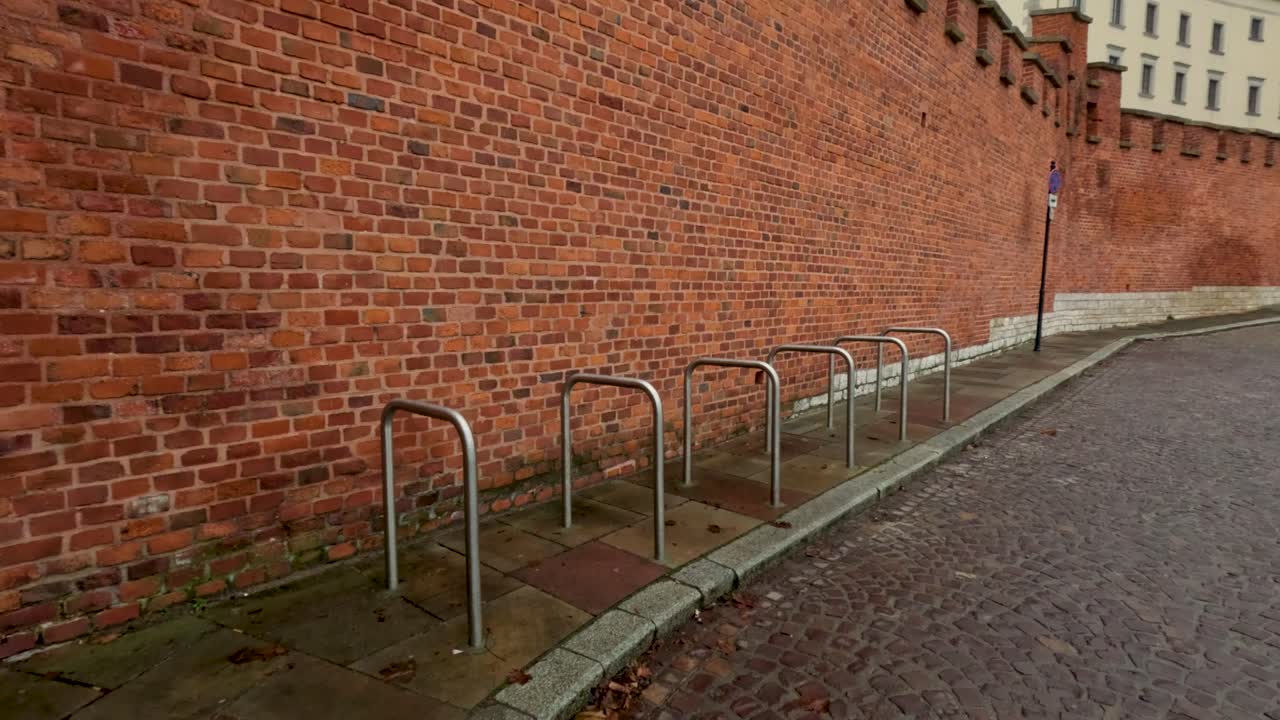 A view of a brick wall with several bike racks set along a curved cobblestone path. Leaves cover parts of the ground, indicating autumn