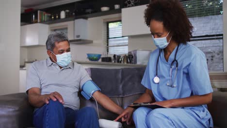 Mixed-race-female-doctor-wearing-mask-and-senior-man-taking-blood-pressure-at-home